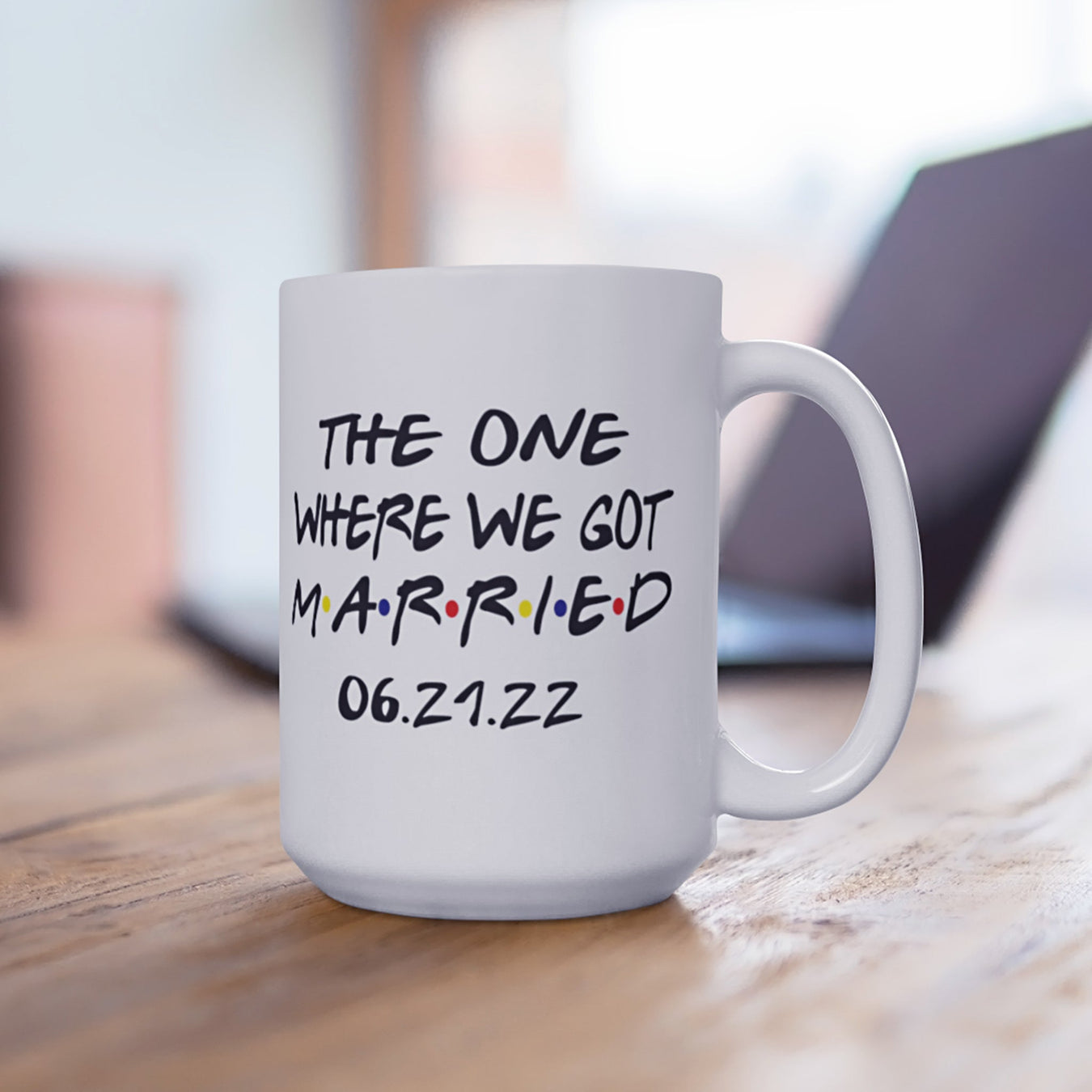 A mug that reads "The One Where We Got Married 06.21.22" sits on a wooden table with a laptop seen in the background.