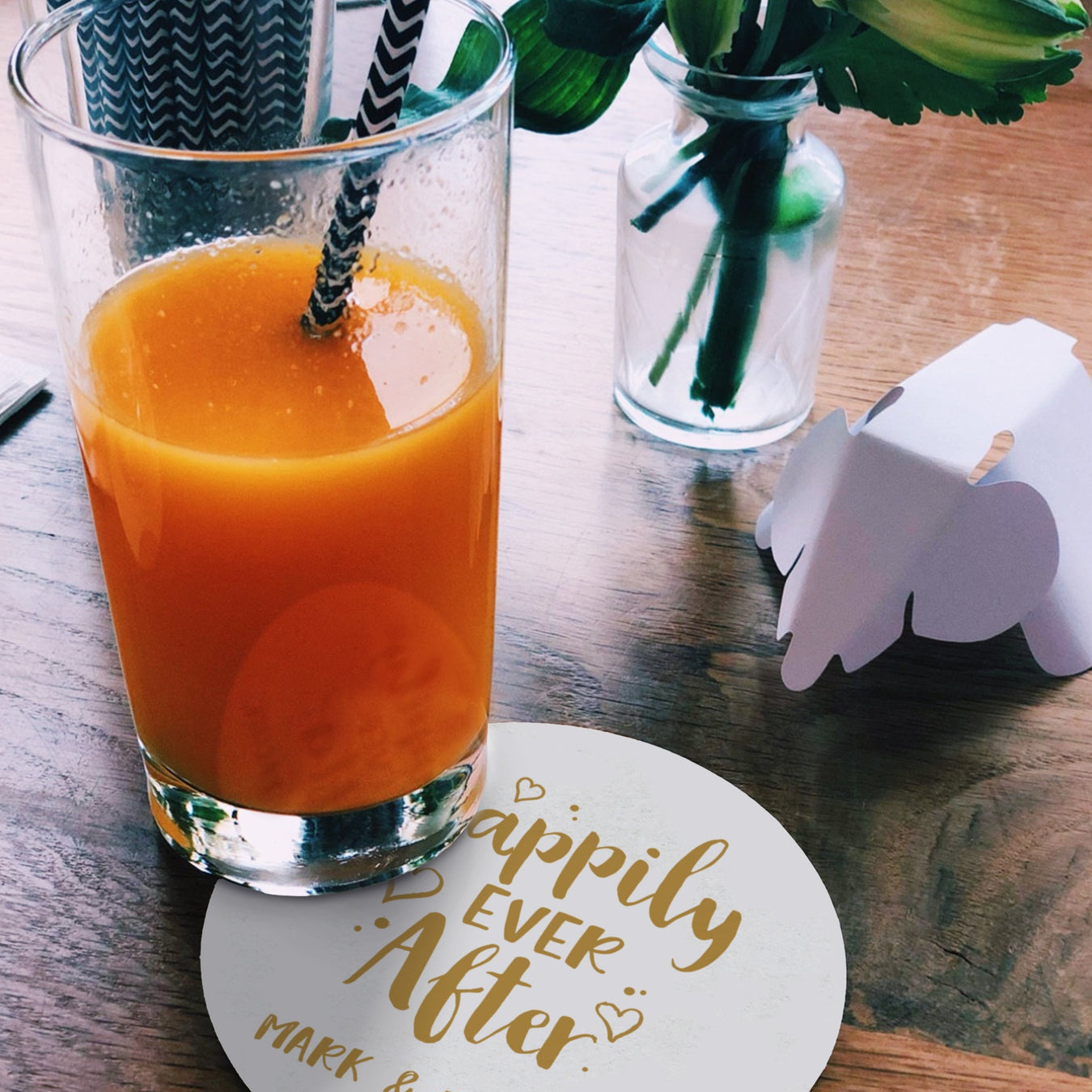 A tablescape is shown with a drink glass on a coaster, a vase, and an origami elephant. The coaster underneatht the glass reads "Happily Ever After Mark & Rachel".