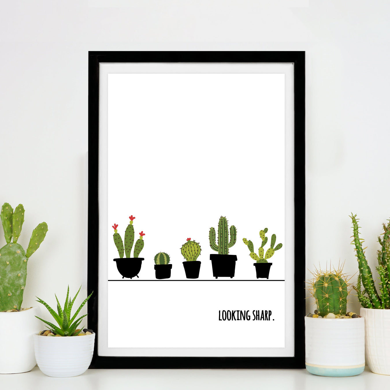 A framed art print that shows different illustrated cacti and the words "Looking Sharp" sits on a white table with small potted succulents and cacti around it.