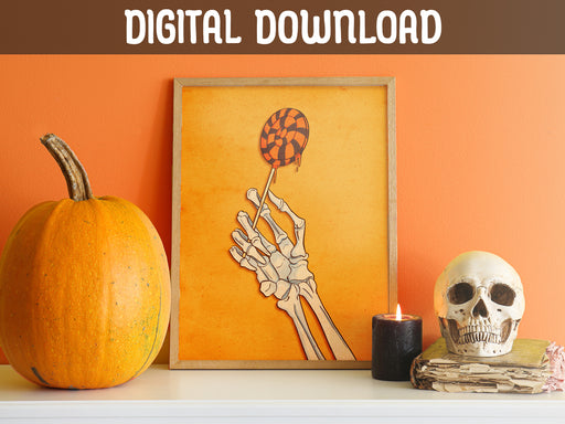 DIGITAL DOWNLOAD: halloween art print of skeleton hand holding a lollipop in a wooden frame in front of an orange wall ontop of a white countertop surrounded by halloween decor such as pumpkins, candles, and a white skull
