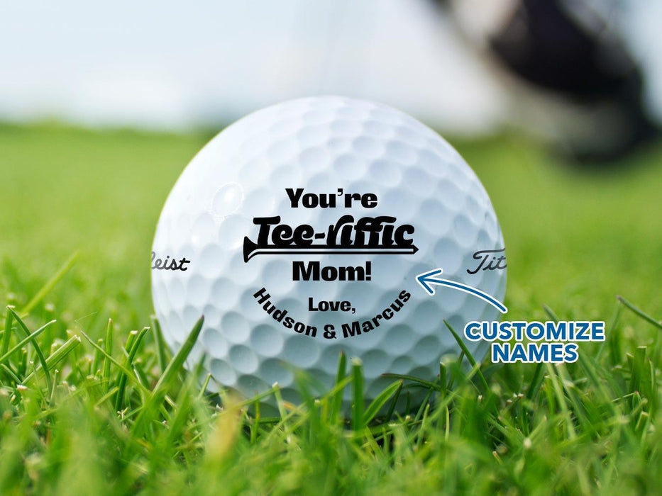 A golf ball with a You're Tee-riffic Mom design printed on it, placed on a grassy field.