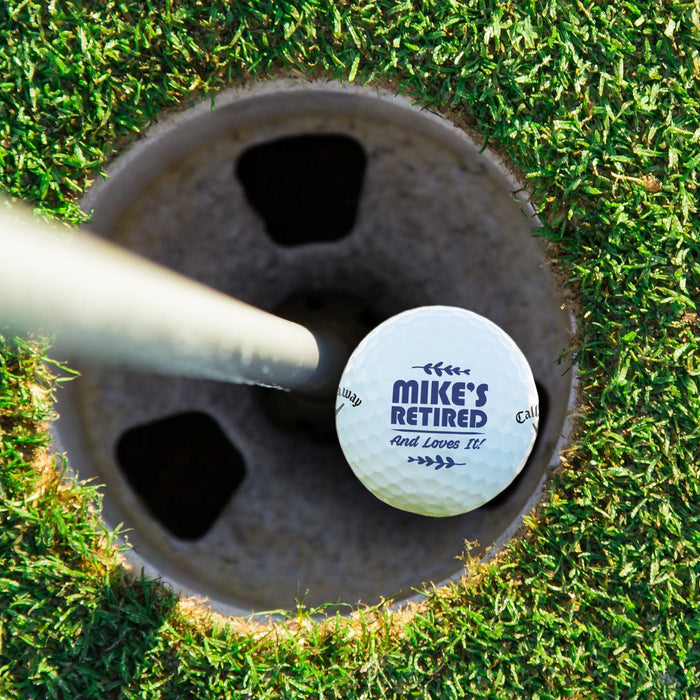 Top view of a personalized retirement golf ball resting in a golf cup. Features the custom "Retired And Loves It" design and personalized name.