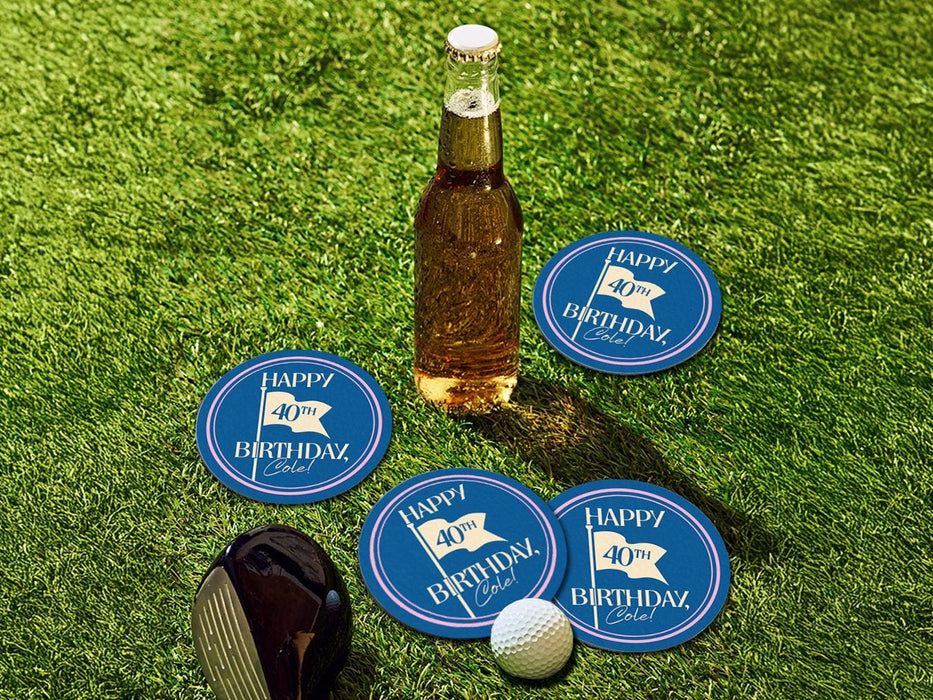 Custom round pulpboard coasters are shown sitting grass with a beer bottle, golf club, and golf ball surrounding them. The custom coasters feature design printed with blue background, pink circular border, and a beige illustrated golf flag.