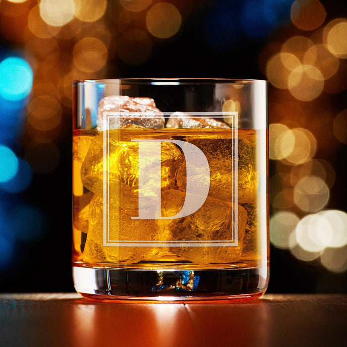 Elegant product photo of the engraved whiskey glass against a soft bokeh background. The framed initial design is centered and clearly visible.