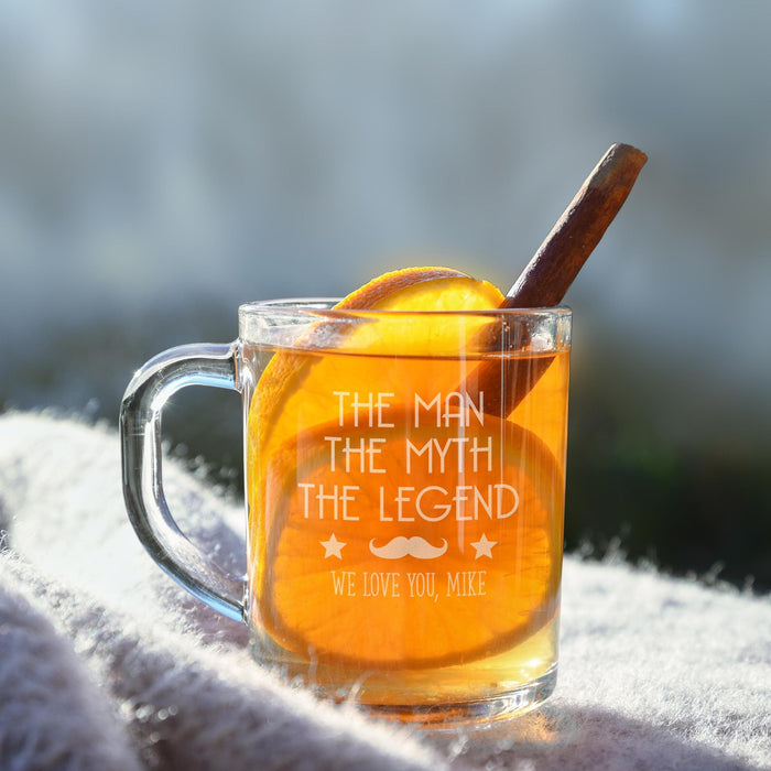 A glass mug filled with a yellow liquid, likely tea, and topped with an orange slice and a cinnamon stick. The mug has a personalized message engraved on it.