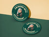 Custom round pulpboard coasters are shown sitting on a yellow and green background. The custom coasters feature design printed with playful &quot;Swinging Into Retirement&quot; text, golf imagery, and custom photo.