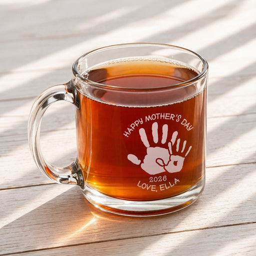 A glass mug filled with a warm beverage, likely tea, sits on a wooden surface. The mug has a handprint design and the text "Happy Mother's Day 2026 Love, Ella" engraved on it.