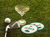 Two custom coasters sit on a grassy golf course next to two martini glasses, a golf club, and a golf ball. The custom coasters features a green border, golf ball, a red flag, and golf hole illustration and bold text across the center that can be customized with a name and lounge name.