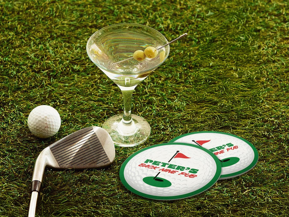 Two custom coasters sit on a grassy golf course next to two martini glasses, a golf club, and a golf ball. The custom coasters features a green border, golf ball, a red flag, and golf hole illustration and bold text across the center that can be customized with a name and lounge name.