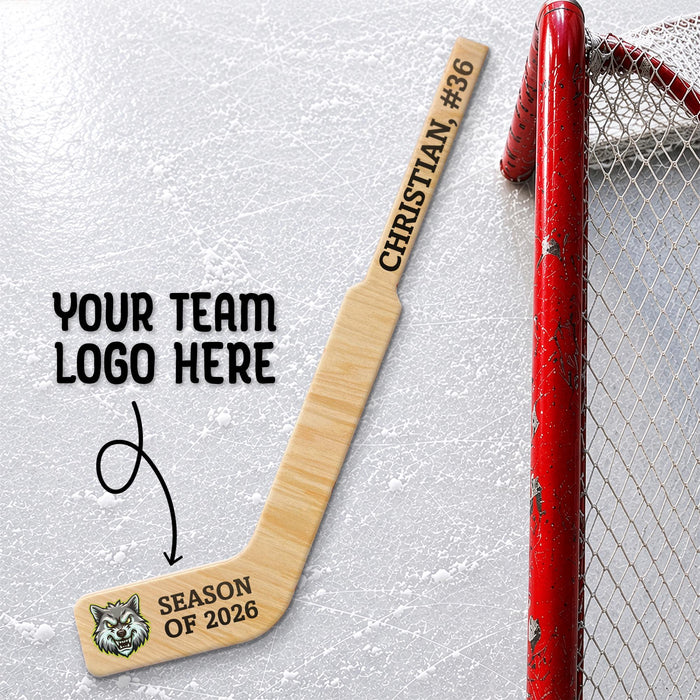 Personalized mini wooden goalie hockey stick featuring custom player name, number, season text, and team logo printed on the blade, displayed beside a hockey net on an ice rink background.