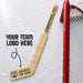 Personalized mini wooden goalie hockey stick featuring custom player name, number, season text, and team logo printed on the blade, displayed beside a hockey net on an ice rink background.