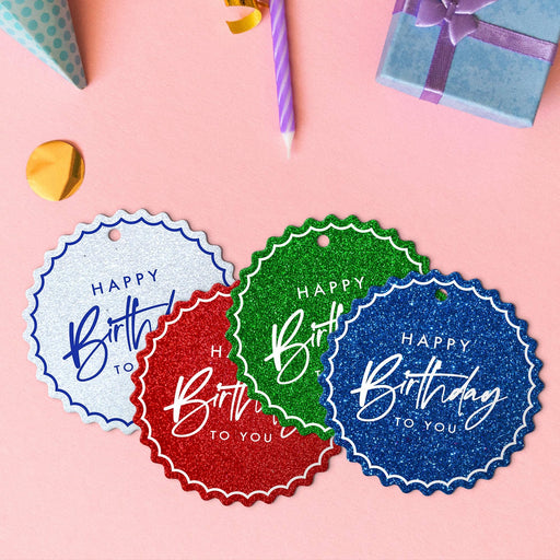 Four birthday hang tags in glitter blue, glitter red, glitter green, and glitter white lying beside colorful gifts and party decorations.