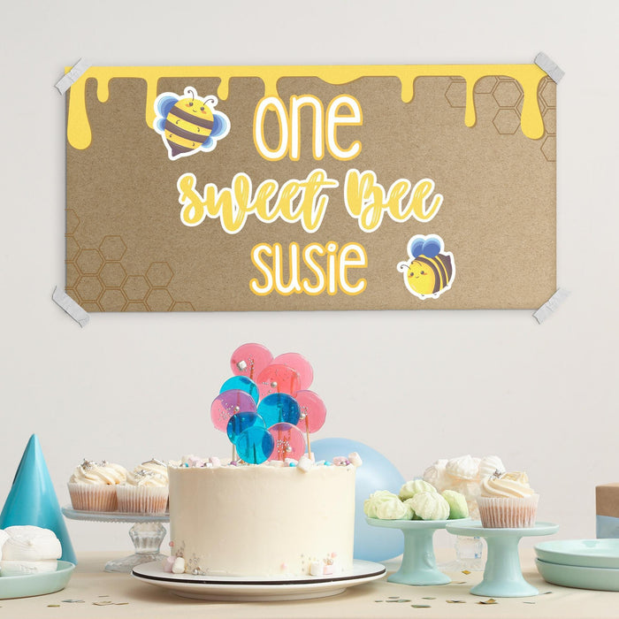 First birthday dessert table setup featuring a custom printed bee themed birthday banner displayed above a birthday cake and party decorations.