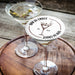 Personalized espresso martini coaster displayed beside two martini glasses on a wooden table, featuring the custom bar name, espresso martini illustration, and established year.