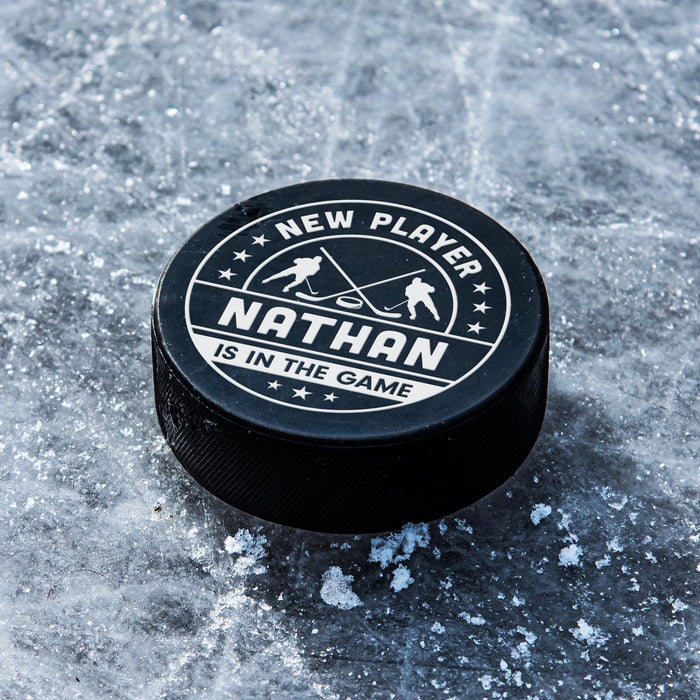 Personalized black hockey puck with a white UV printed “New Player” baby birth announcement design resting on ice.