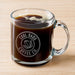 A glass mug filled with a dark liquid, likely coffee, with foam on top. The mug has an engraving on it that says "Cool Dads Coffee Co."