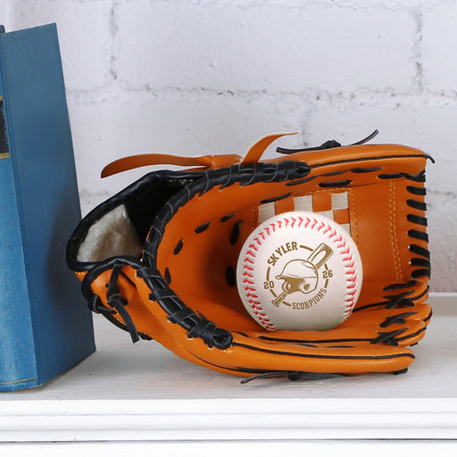 A baseball glove with a personalized baseball inside it, placed on a white surface next to a blue book. A baseball is featuring an engraved design with a name, a year, a team name and a baseball helmet and bat graphic.