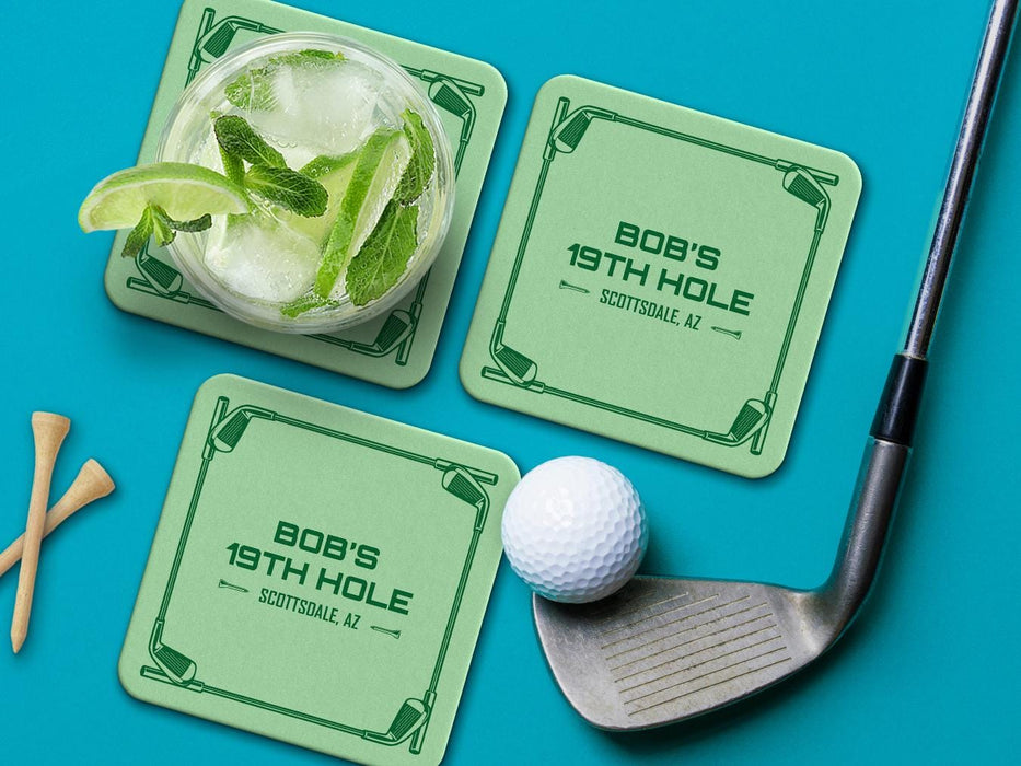 Custom square pulpboard coasters are shown sitting on a blue surface. Golf tees, a golf ball, and a golf club sit beside them. One coaster has a cocktail on top of it. The custom coasters feature a green golf bar design with interlocking golf clubs used as a border. The bar name and location can be customized.
