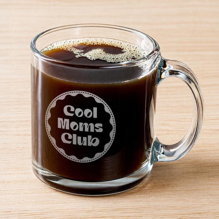 A glass mug filled with a dark liquid, likely coffee, with foam on top. The mug has a handle and an engraving on it that says "Cool Moms Club".