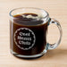 A glass mug filled with a dark liquid, likely coffee, with foam on top. The mug has a handle and an engraving on it that says "Cool Moms Club".