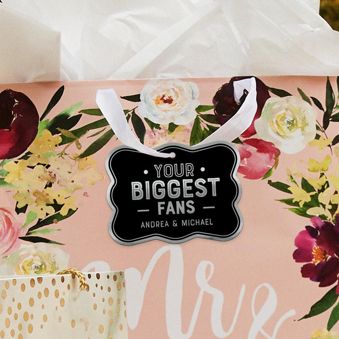 Floral wedding-themed flat lay featuring a personalized “Your Biggest Fans” foil wedding gift tag in black and metallic silver. Styled with romantic flowers and wedding décor accents for elegant gifting inspiration.