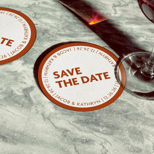 Personalized Save the Date coaster displayed beside a wine glass. Features a bold circular border design with custom names and wedding date.