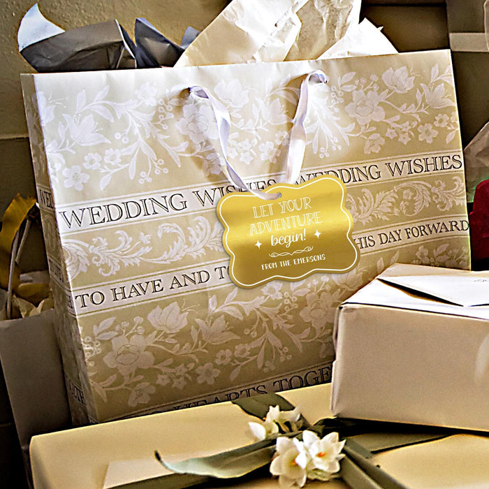 Gold foil “Let Your Adventure Begin” wedding gift tag attached to elegant wedding gift packaging. Styled with wrapped presents and neutral wedding décor for gifting inspiration.
