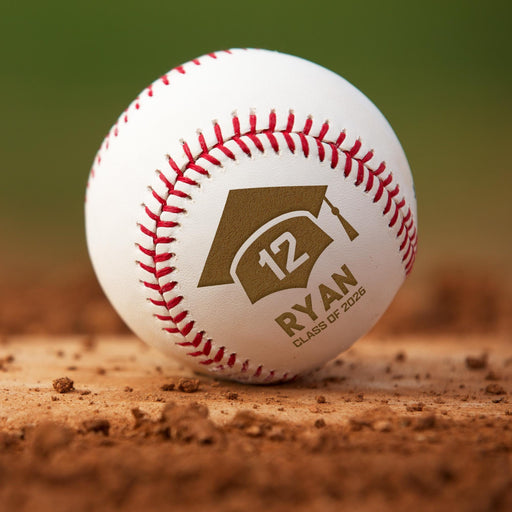 A personalized baseball with red stitching and a graduation cap logo engraved on it, placed on a dirty base surface.