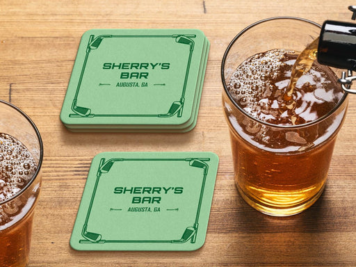 Custom square pulpboard coasters are shown sitting on a wooden table next to two drink glasses. The custom coasters feature a green golf bar design with interlocking golf clubs used as a border. The bar name and location can be customized.