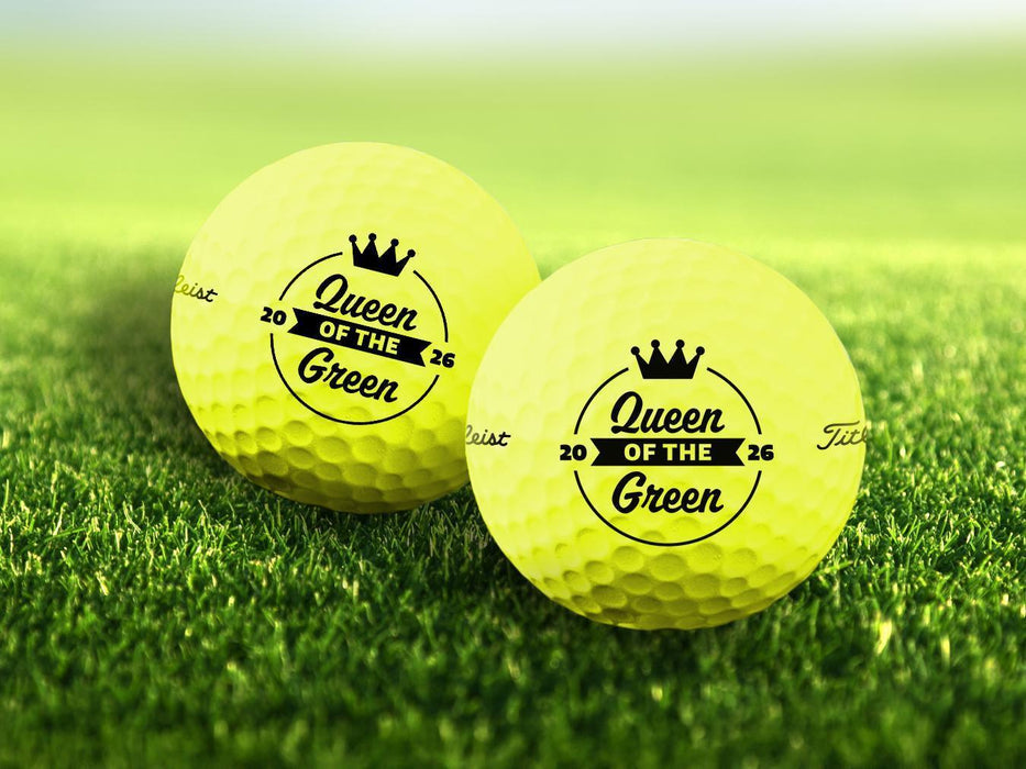 Two personalized yellow golf balls with a Queen of the Green design printed on them, placed on a grassy field.