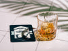 Custom square pulpboard coaster are shown laid out on marble floor. A whiskey glass is shown next to the coaster. The coaster features design with an illustration of a golf ball and with club behind it and custom text.