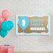 Personalized blue first birthday banner hanging on a white brick wall featuring balloon artwork and custom printed name design.
