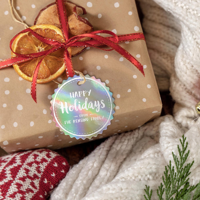 Personalized Happy Holidays round hang tag tied to a kraft paper gift with red ribbon, dried orange slices, greenery, and a cozy knit background.