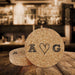 Personalized cork coasters with hand-drawn heart initials displayed in a rustic bar setting.