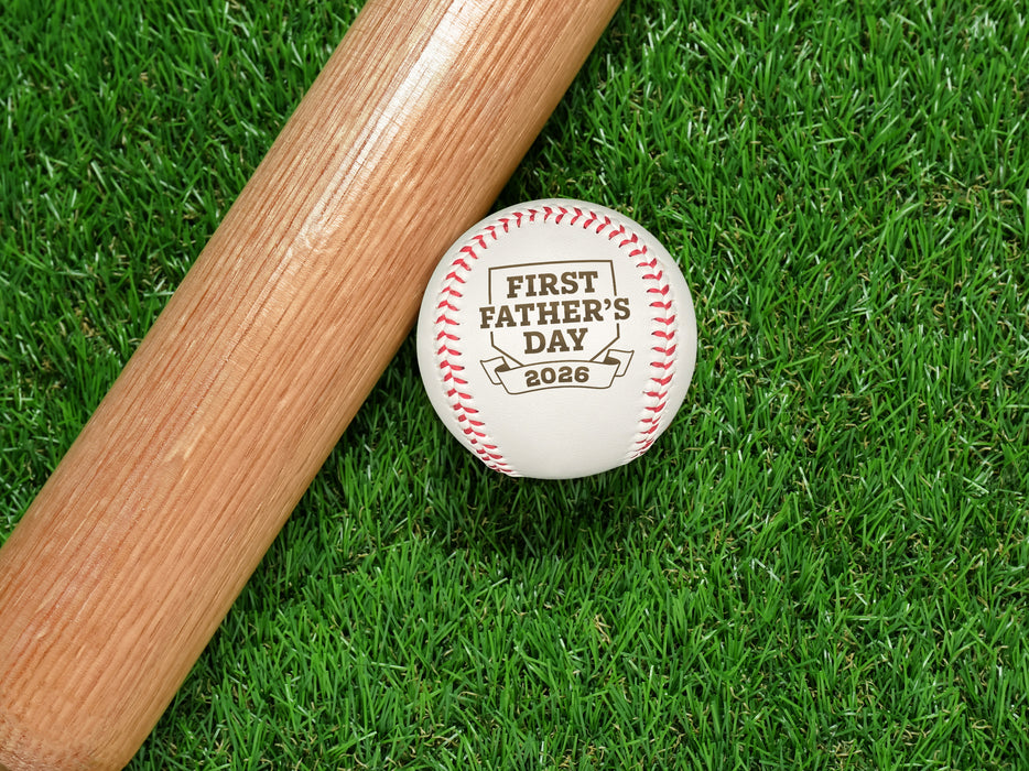 Baseball sits on grass next to baseball bat. Baseball has First Father's Day design engraved on it.