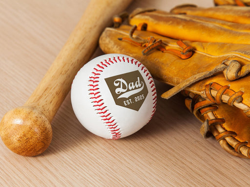 Baseball, wooden bat, and glove sits on wooden surface. Baseball has Dad Established Date design engraved on it.