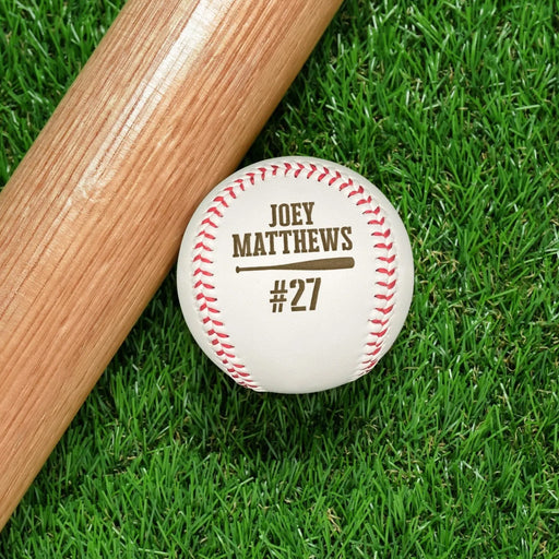 An engraved baseball and wooden baseball bat sits on a green grassy background. The engraved design features the player's name and jersey number along with a baseball bat graphic.