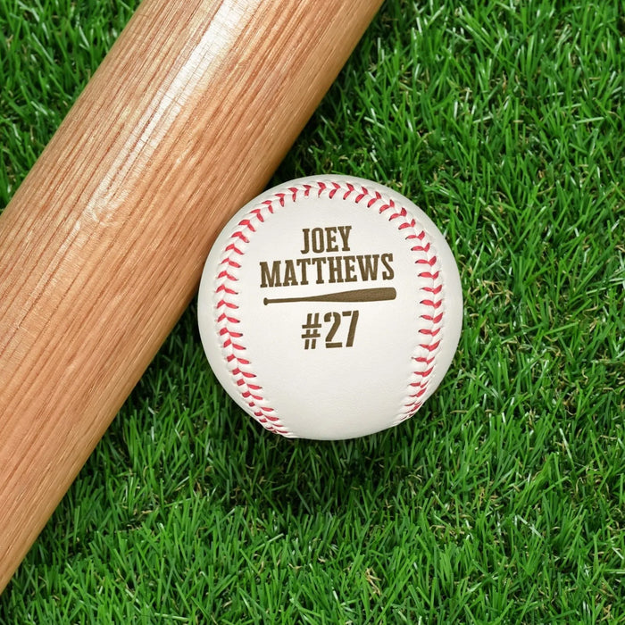 An engraved baseball and wooden baseball bat sits on a green grassy background. The engraved design features the player's name and jersey number along with a baseball bat graphic.