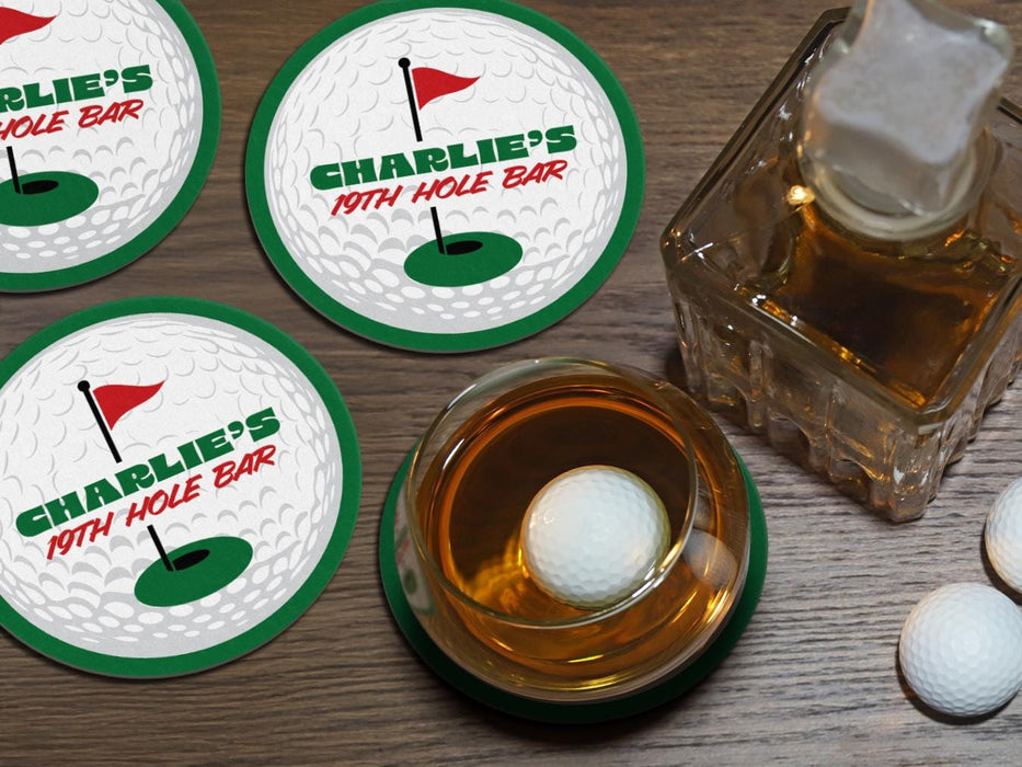 A full bourbon glass is shown next to a decanter while sitting on a wooden table. A golf ball rests in the glass. Custom coasters and golf balls can be seen on the table as well. A custom coaster is also placed underneath the glass. The custom coasters features a green border, golf ball, a red flag, and golf hole illustration and bold text across the center that can be customized with a name and lounge name.