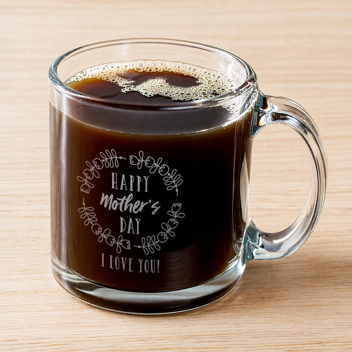 A glass mug filled with a dark liquid, likely coffee, with foam on top. The mug has a message engraved on it that says "Happy Mother's Day I Love You!"