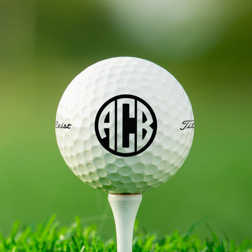 single white titleist golf ball on white golf tee with custom personalized black circle monogram ABC printed on it against grass golf field background
