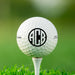 single white titleist golf ball on white golf tee with custom personalized black circle monogram ABC printed on it against grass golf field background