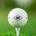 single white titleist golf ball on white golf tee with custom black diamond monogram design ABC printed on it against grass golf field background