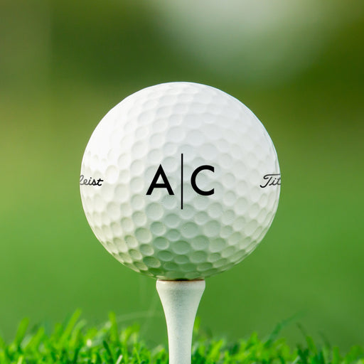 single white titleist golf ball with custom personalized black line monogram printed design on white golf tee against grass golf course background