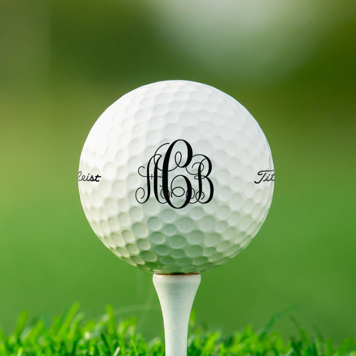 single white titleist golf ball with custom personalized black scroll monogram printed design on white golf tee against a grass golf course background
