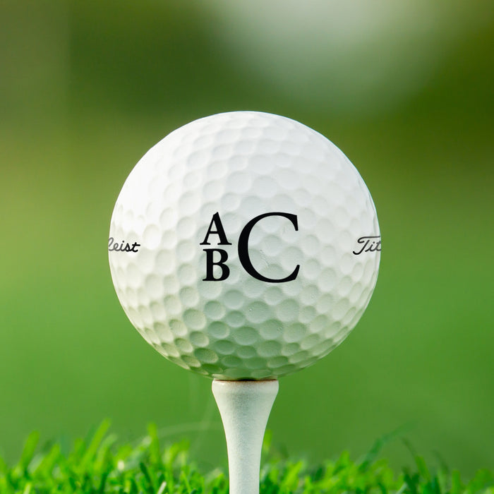 single white titleist golf ball with custom personalized black stacked monogram printed design on white golf tee on top of golf course grass background 