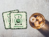 Two coasters sit on stone table with whiskey glass next to them. The coaster features design with illustrated golf ball, club, and flag on top with wreath surrounding it, argyle background pattern, and custom text.