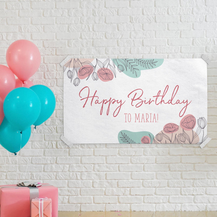 Floral celebration banner displayed on a white brick wall with colorful balloons and party decorations.
