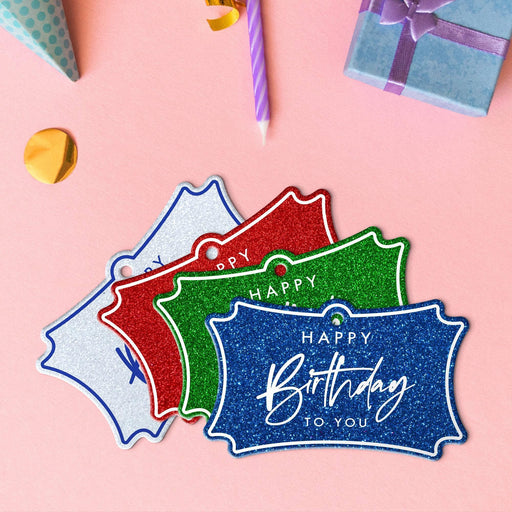Four elegant script birthday hang tags in glitter red, glitter blue, glitter green, and glitter white lying beside colorful gifts and party decorations.