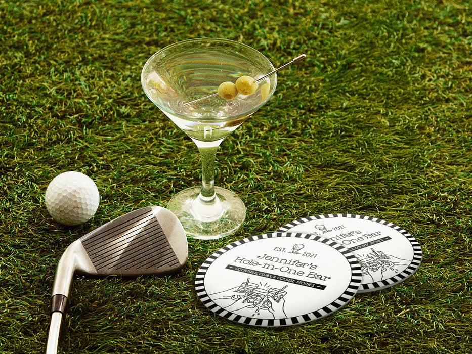 Two custom coasters sit on a grassy golf course next to two martini glasses, a golf club, and a golf ball. The custom coasters feature a golf club lounge design with an illustration of a golf ball on a tee and people cheersing drinks together along with a striped border. The established year, name, lounge name, and tagline can be customized.